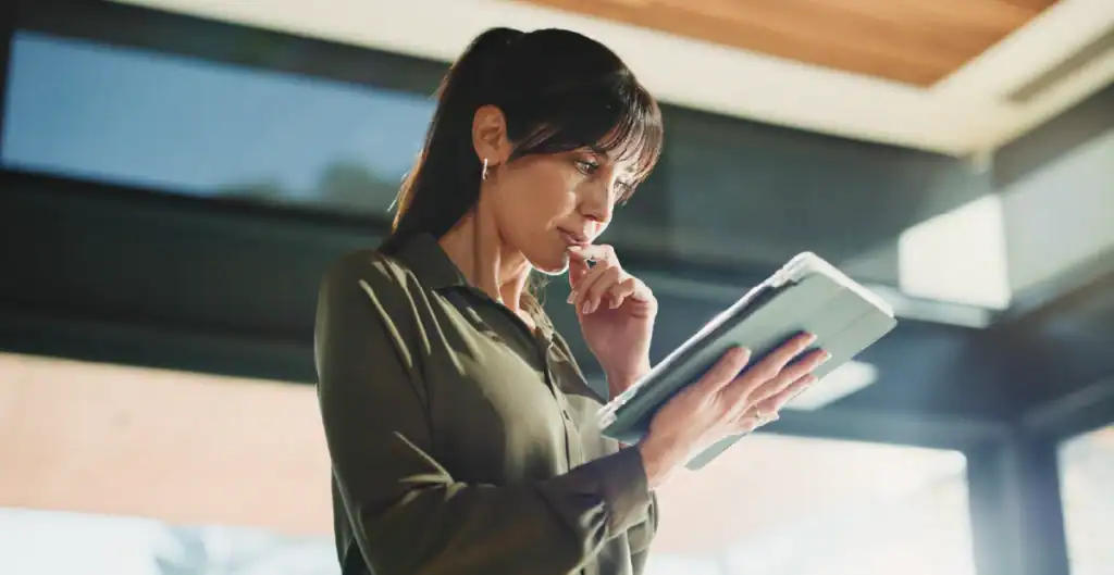 A woman in a green blouse stands indoors, looking thoughtfully at a tablet while holding her glasses to her lips. Sunlight enters through large windows behind her.
