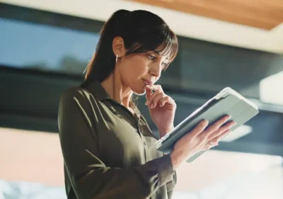 A woman in a green blouse stands indoors, looking thoughtfully at a tablet while holding her glasses to her lips. Sunlight enters through large windows behind her.