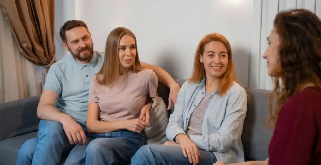 Four adults sit on a couch, smiling and talking. Three face forward, one faces them. They appear relaxed, casually dressed, and engaged in friendly conversation in a cozy indoor setting.