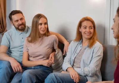 Four adults sit on a couch, smiling and talking. Three face forward, one faces them. They appear relaxed, casually dressed, and engaged in friendly conversation in a cozy indoor setting.